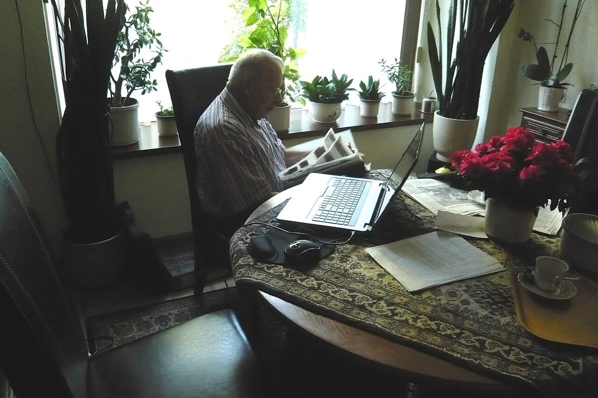 Older man with white hair using a laptop at a desk with plants and flowers, browsing Facebook groups for photographers