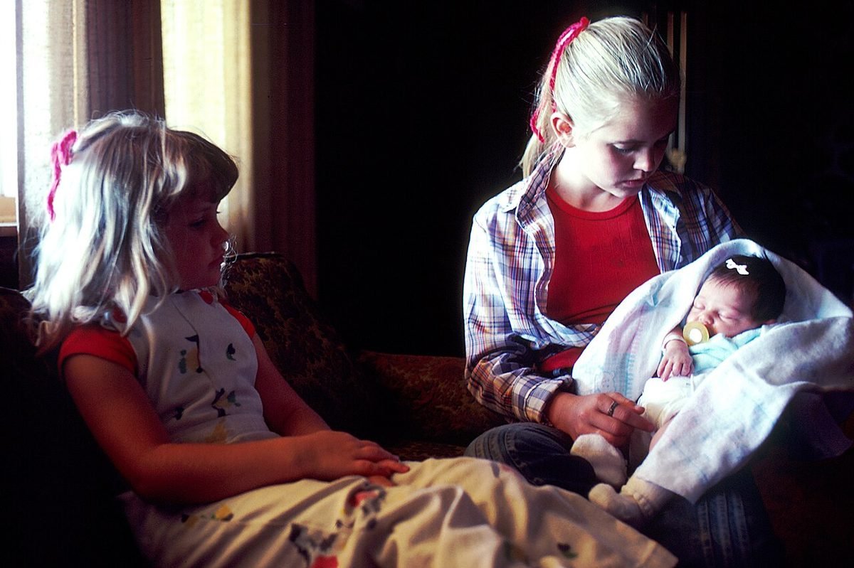 Two blonde girls sit close on an indoor sofa; one cradles a swaddled newborn while the other looks on.