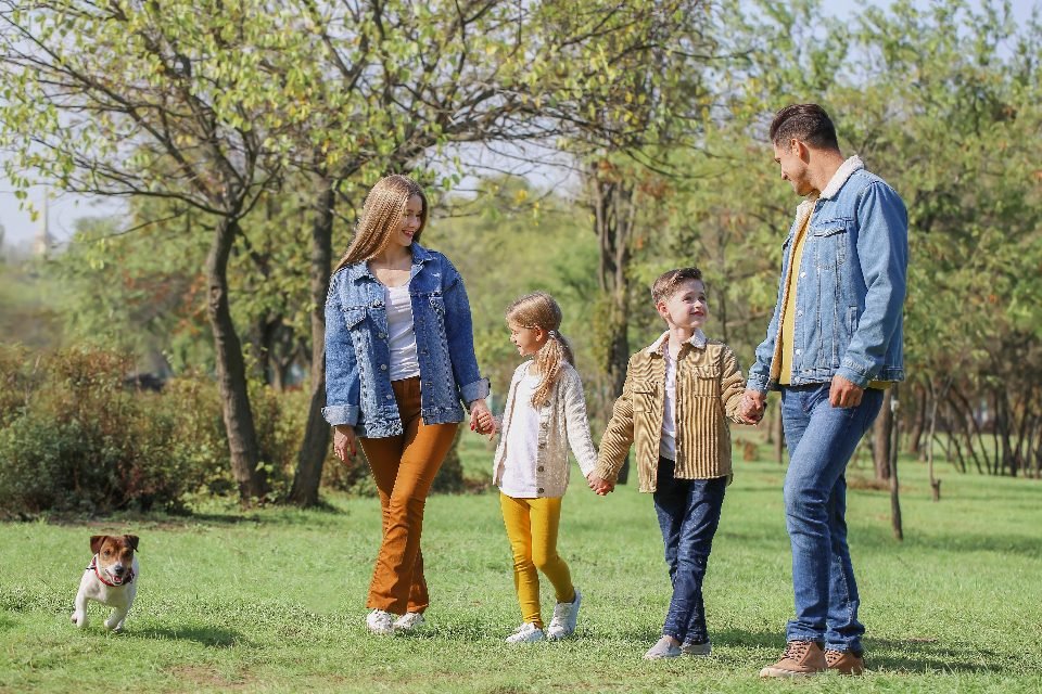 Family photo posing ideas: a family of four holding hands while walking across a sunny park lawn with a small dog.