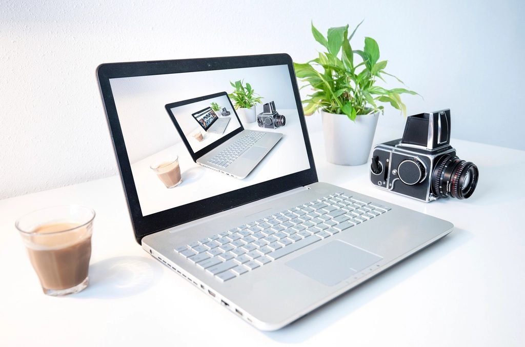 Laptop on a white desk beside a glass of coffee, a potted plant and a medium-format film camera.
