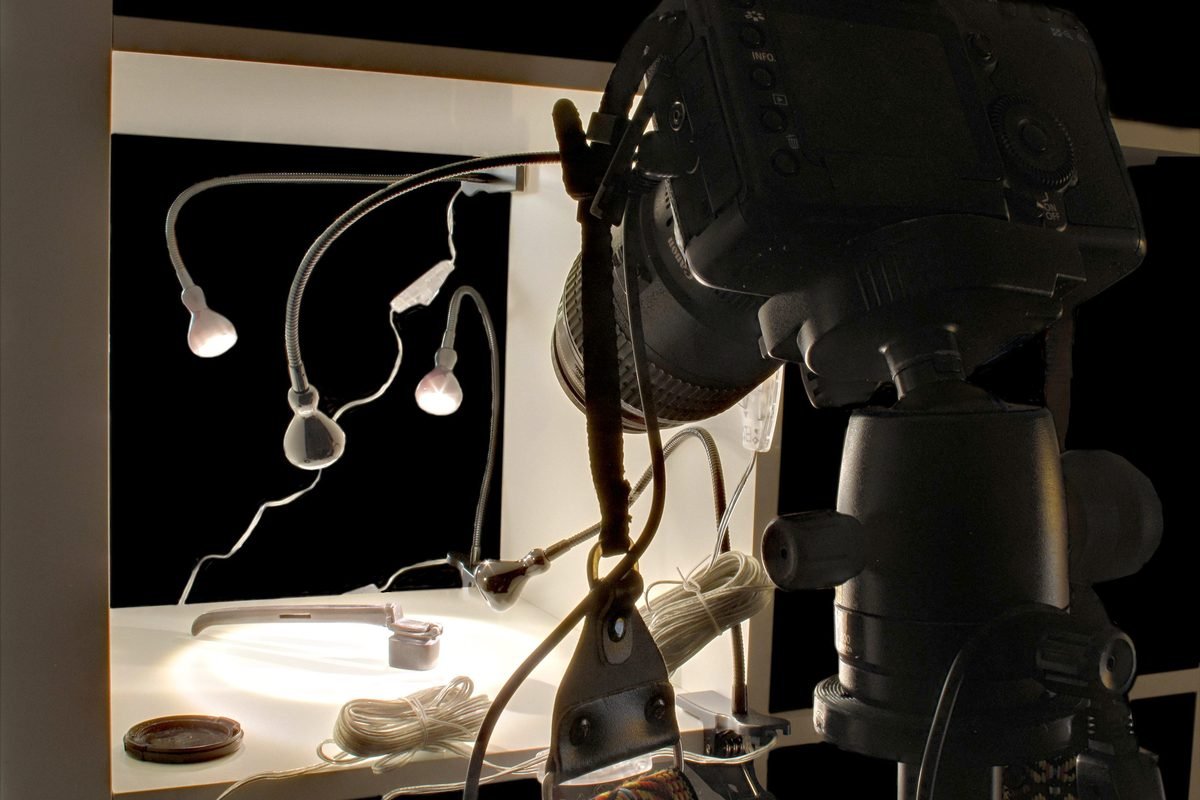 Camera on a tripod aimed at a small tabletop studio with a black backdrop, flexible lights, and cables.