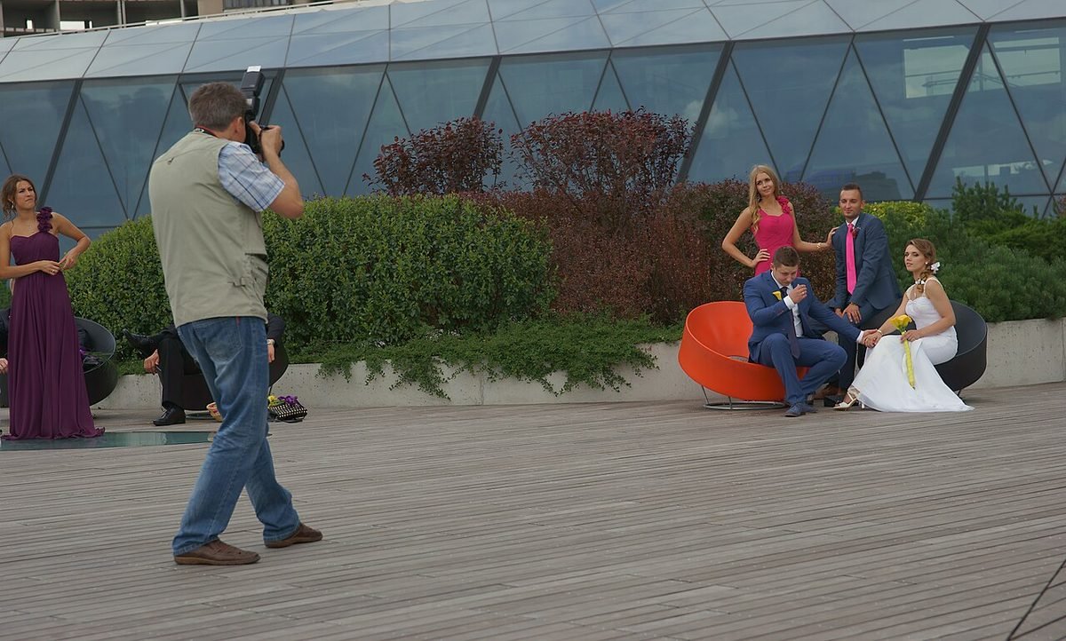 Wedding photographer in a vest shooting a bride and groom seated on an orange chair on an outdoor wooden deck