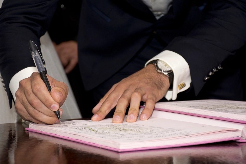 Man in a dark suit with a wristwatch signing a document in a pink folder on a glossy table, holding a black pen.