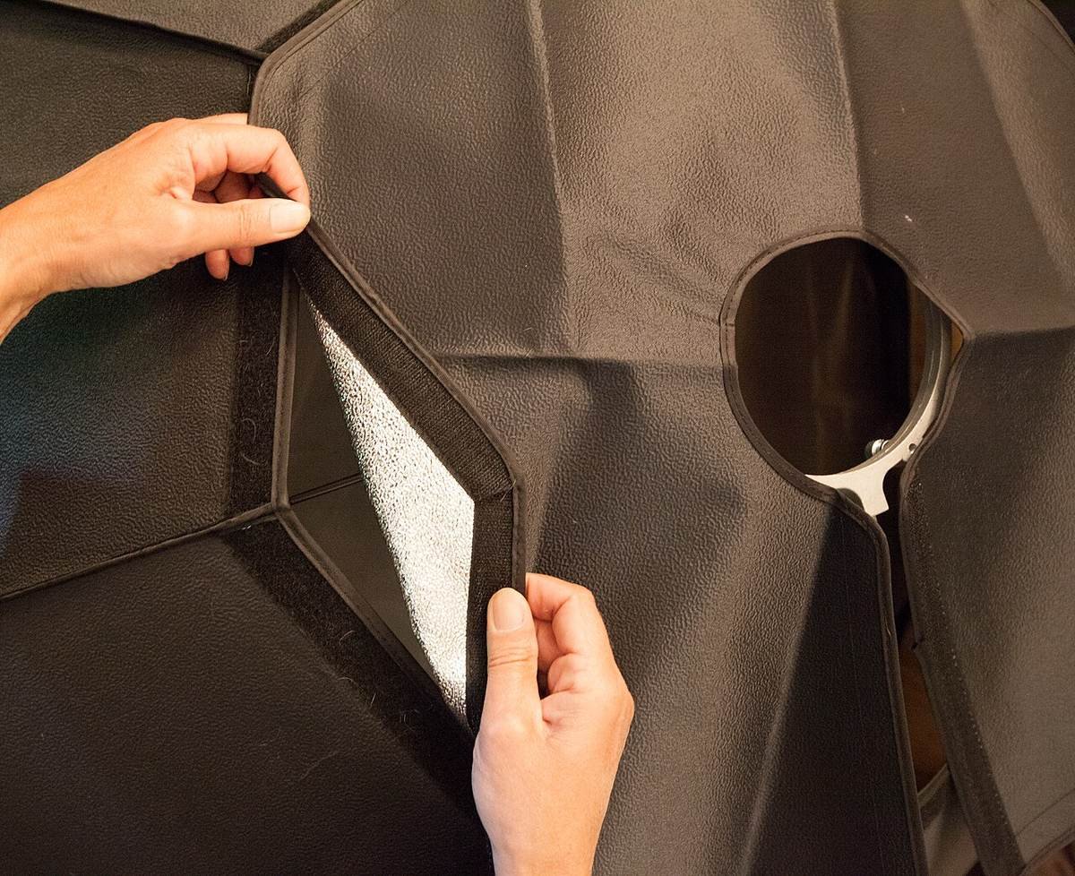 Hands adjusting the black velcro flap of an octagonal studio softbox with a silver interior and a light mount.