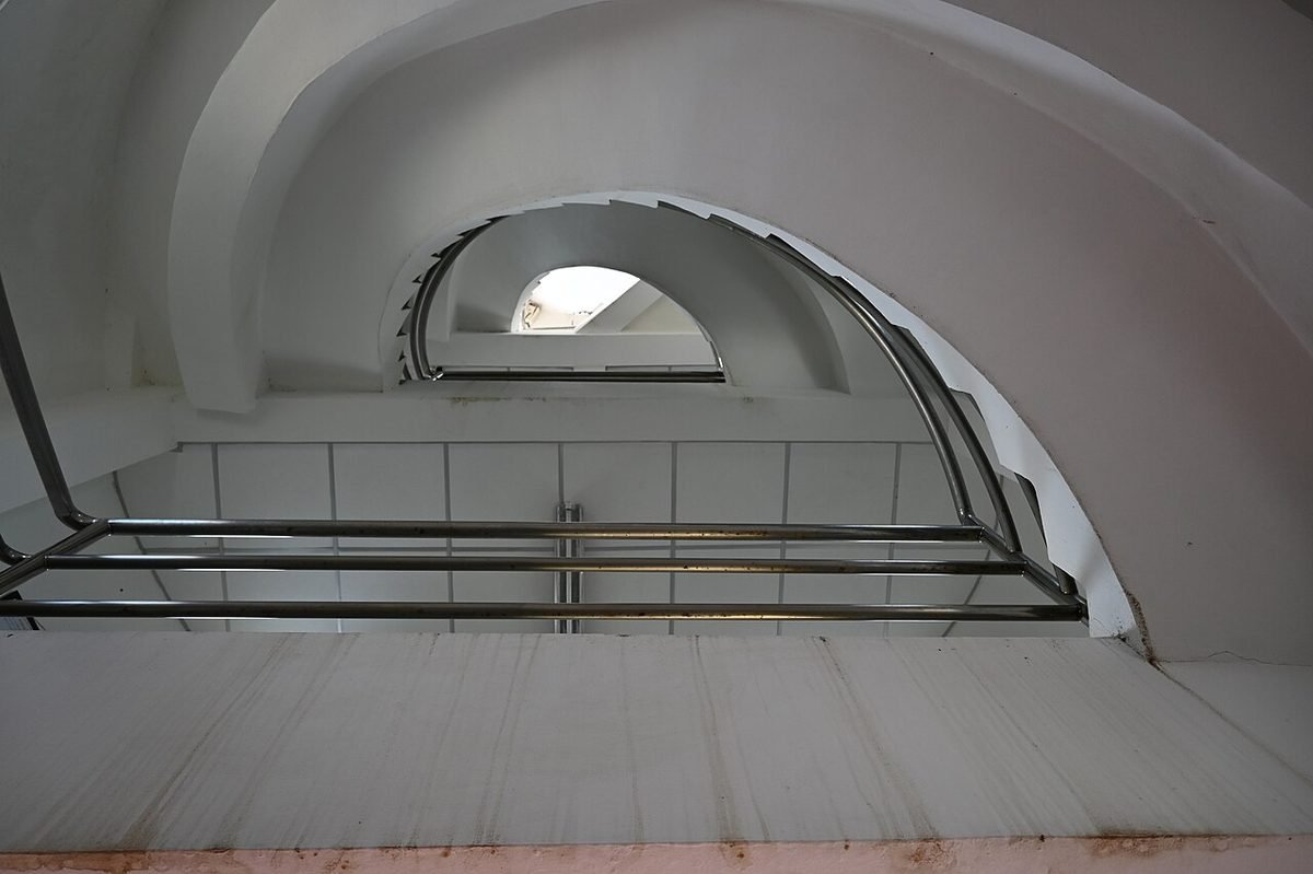 Curved white staircase with chrome handrails leading up toward a bright arched opening in a whitewashed vaulted ceiling.